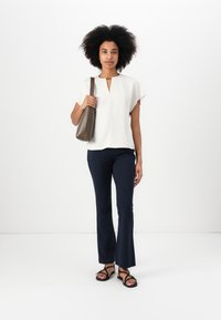 Haut blanc à manches courtes avec un décolleté en trou de serrure, associé à un pantalon ajusté bleu marine et des sandales noires. Le modèle porte un sac à épaule marron.