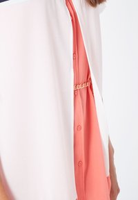 Blusa con un frente de botones coral, con una superposición blanca y transparente. Un detalle de cadena plateada acentúa la costura lateral.