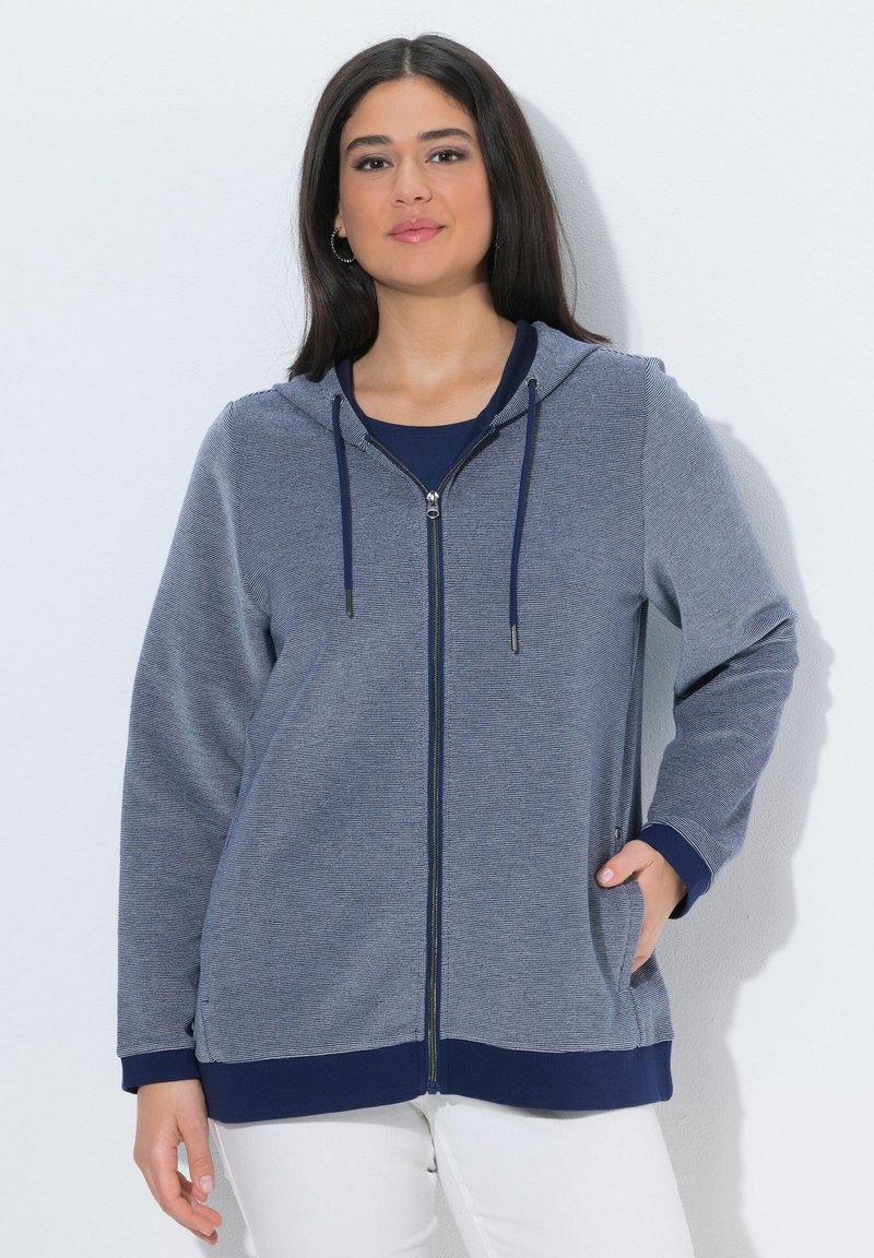 Mujer con cabello largo y oscuro, vistiendo una sudadera azul con cremallera sobre una camiseta azul marino y pantalones blancos, de pie con una mano en el bolsillo.