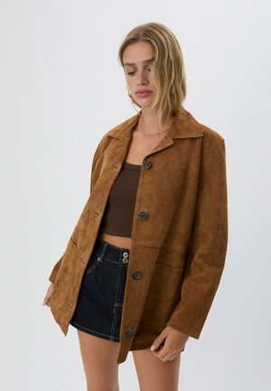Femme portant une veste boutonnée en daim marron, un crop top marron foncé et une minijupe en denim foncé sur fond clair, regardant vers le bas.
