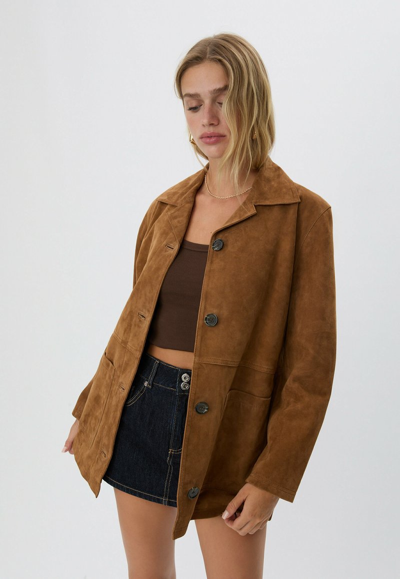 Kvinna i brun mockajacka med knappar, mörkbrun crop top och mörk denim mini kjol framför ljus bakgrund, tittar ner.