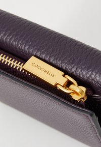 Purple leather wallet with a textured finish, featuring a golden zipper with a brand tag that reads "Coccinelle."