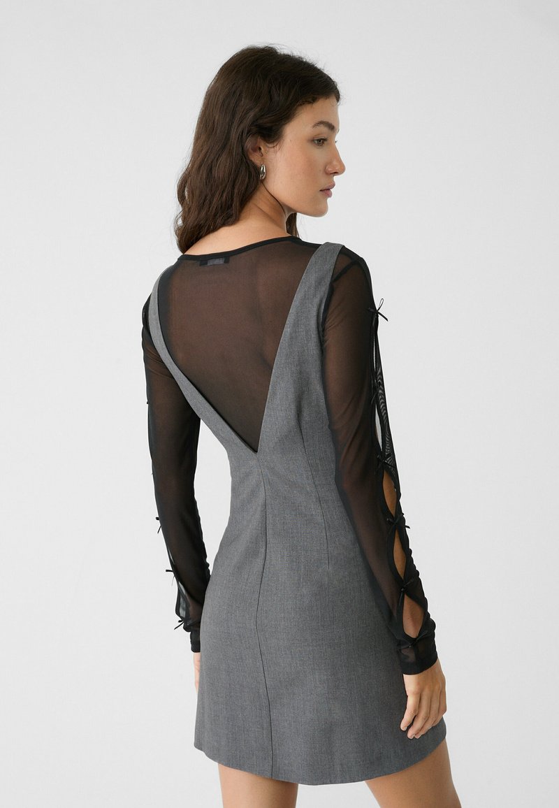 Abito grigio con corpetto aderente, scollatura a V profonda sul retro e maniche lunghe trasparenti nere con dettagli di tagli e lacci ai polsi.