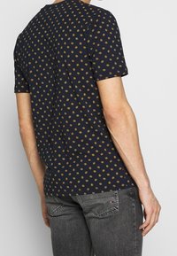 Hombre vestido con una camisa de manga corta negra con pequeños patrones florales amarillos y jeans negros desgastados, visto desde atrás frente a un fondo liso.