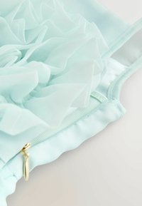 Heleda sinine pluus, millel on kihilised volangid õhukesest kangast. Küljel kuldsete detaile sisaldav lukuga sulgemine, pehme tekstuur.