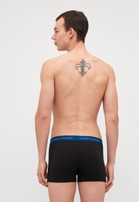 Musta bokseriláhke tummuhi tummuja elastisella vyötärönauhalla, johon on kirjailtu "TOMMY HILFIGER" -logo. Mallilla on tatuointi selässään, hän katsoo poispäin.
