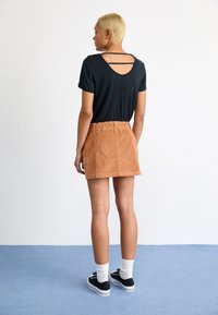Zwarte t-shirt met een grote uitsnijding op de rug en een tan corduroy mini-jurk. Model draagt zwarte sneakers en witte sokken, staande op een blauwe ondergrond.