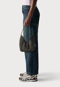 Sac en cuir gris foncé avec une forme de pochette distinctive, doté d'une fermeture à boucle. Porté avec un jean droit bleu et des baskets en matériaux mélangés.