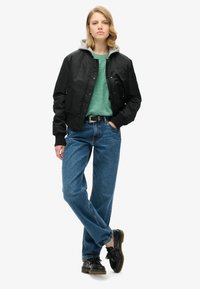 Sort bomberjakke med grå hætte, grøn skjorte og blå jeans. Modellen har sorte sko med kvaster på og står foran en hvid baggrund.