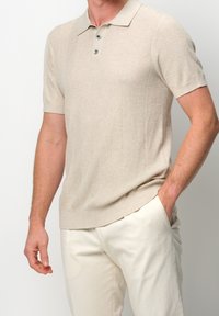 Polo de manga corta en color beige con un patrón texturizado, tapeta de tres botones y una tela suave de punto fino. Combinado con pantalones de color claro.