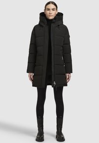 Melna puffer jaka ar kapuci, ar quilted daļām, priekšējo rāvējslēdzēju un sānu kabatām. Valkāta virs melna apģērba un legingiem.