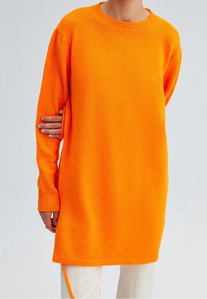 Person iført en knaldorange langærmet sweaterkjole over lyse bukser, stående mod en ensfarvet lysegrå baggrund.