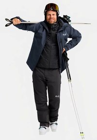 Ski-Bekleidung: navyblaue Jacke mit durchgehendem Reißverschluss, schwarze isolierte Weste, schwarze Hose und weiße Skistiefel. Zubehör: Skistöcke und Skibrille.
