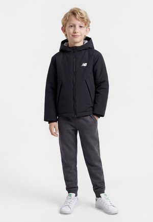 Ragazzo con capelli biondi che indossa una giacca nera New Balance, pantaloni grigio scuro e scarpe da ginnastica bianche, in piedi con una mano in tasca.