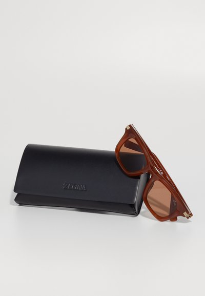 Brown sunglasses with a square frame and brown tinted lenses, resting on a black leather case embossed with "ZEGNA."