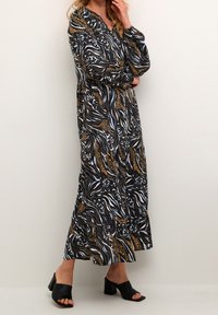 Robe longue à manches longues, avec un motif tourbillonnant en noir, blanc, bleu et doré. Associée à des mules à talons noires.