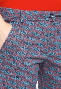 Kalhoty se vzorem s modro-červeným opakujícím se designem a zakřivenými motivy, vyrobené z lehké tkaniny. Obsahují přední kapsu a detail s knoflíkem.