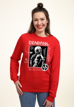 Vrouw glimlacht en draagt een rode Deadpool-trui met stripfiguur grafiek en tekst, gecombineerd met een blauwe spijkerbroek tegen een effen achtergrond.