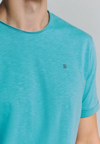 T-shirt met korte mouwen en ronde hals in licht teal, met een gestructureerde stof, omgerolde mouwen en een klein zwart logo op de borst.