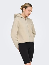 Beige fleece cropped hoodie with hood and front zipper. Textured fabric, paired with black ribbed bicycle shorts.