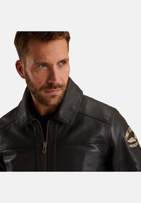 Čierna kožená bunda na zips s širokým golierom. Obsahuje šitie na pleciach a vyšívanú nášivku na pravej rukáve.