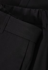 Pantalon en tissu noir présentant une texture lisse, une ceinture plate et des plis latéraux, offrant un design simple et épuré.