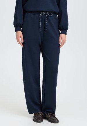 Pantalon bleu marine à cordon porté avec un haut assorti et des chaussures décontractées marron foncé, présenté de la moitié du torse jusqu'aux pieds sur un fond clair uni.