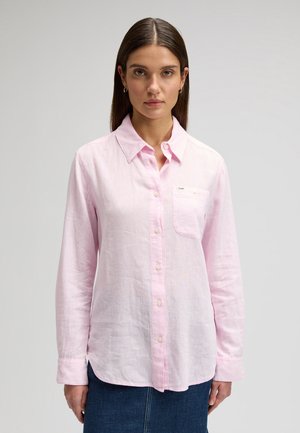 Junge Frau mit glatten braunen Haaren trägt ein hellrosa Hemd mit Knöpfen und einen dunkelblauen Rock, steht vor einem schlichten hellgrauen Hintergrund.