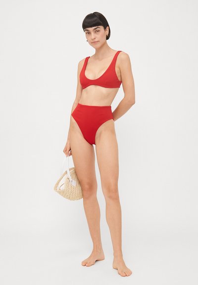 Bikini rosso con una parte superiore scollata e pantaloni a vita alta. Materiale liscio, design minimale, abbinato a una piccola borsa intrecciata.