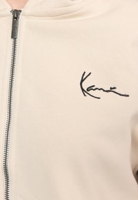 Beige zip-hoodie gemaakt van zachte stof, met een zwart geborduurd logo op de linkerbovenborst en een zilveren rits aan de voorkant.