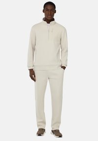 Felpa beige chiaro a mezza zip con tasca sul petto, abbinata a pantaloni beige coordinati e scarpe sportive scure. Tessuto morbido e liscio.