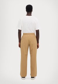 Ein Mann steht mit dem Rücken zur Kamera, trägt ein weißes T-Shirt mit einer schwachen Nummer 7 auf dem Rücken, beige Hose mit Gesäßtaschen und weiße Turnschuhe.