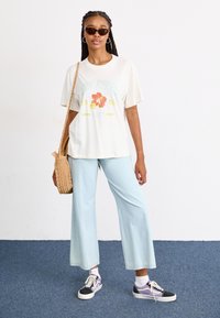 T-shirt beige clair avec des graphiques floraux verts et orange, associé à un jean large bleu clair et des baskets noires avec des accents violets.