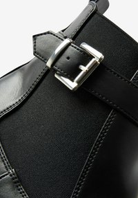 Bottine noire à la cheville avec extérieur en cuir brillant et panneau élastique texturé, agrémentée d'une boucle argentée.