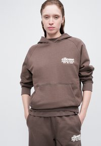 Kobieta o jasnej karnacji z mokrymi włosami, ubrana w brązowy hoodie ellesse i dopasowane dresy, stoi z rękami w kieszeniach na jednolitym tle.