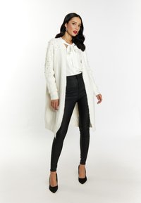 Cardigan alb tricotat cu accente de perle, purtat peste o bluză albă cu fundă. Pantaloni negri din piele ecologică și tocuri stiletto negre completează ținuta.