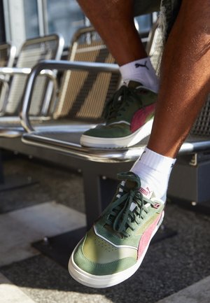 Baskets vertes et bourgogne avec une finition en daim, semelle en caoutchouc blanc et lacets ; portées avec des chaussettes blanches sur un banc métallique.