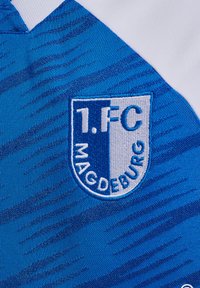 Maillot de sport bleu et blanc avec le logo brodé du 1. FC Magdeburg. Le tissu a une texture lisse avec un motif en vagues.