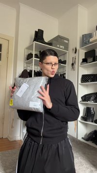 Sivi dukserica u prozirnom pakiranju s vidljivim oznakama. Model nosi crnu duksericu na patent zatvarač i hlače, pozirajući u modernom unutarnjem okruženju.