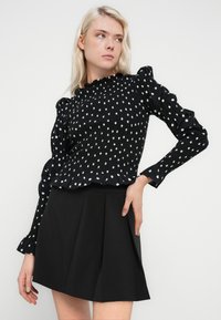 Bluza neagră cu buline albe are un design cu inserții smocate, guler cu volane și mâneci bufante, asortată cu o fustă plisată neagră.