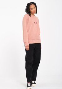 Rosa Sweatshirt mit Rundhalsausschnitt und sichtbaren Nahtdetails, kombiniert mit schwarzen weit geschnittenen Jeans und schwarzen Turnschuhen mit weißen Schnürsenkeln.