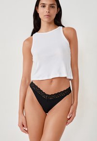 Weißes, kurzes Tanktop und schwarzer Slip mit Spitzenbesatz, der eine sanfte, glatte Textur und aufwendige Spitzenmuster entlang des Taillenbunds aufweist.