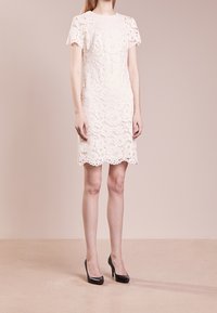 Kvinna som bär en vit spetsklänning med korta ärmar i knä längd, tillsammans med svarta högklackade skor, står på ett beige golv.