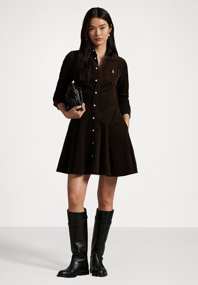 Brown corduroy shirt dress with a fitted bodice, flared skirt, long sleeves, and white buttons. Paired with knee-high black boots.