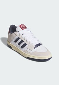 White sneaker with beige suede accents, three black stripes on the side, round red logo on the tongue, and textured rubber outsole.