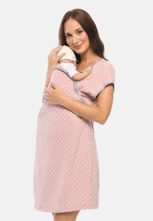 Woman in pink polka-dot dress holding a sleeping baby wearing a white hat, cradled close to her chest against a plain background.