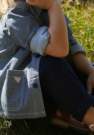Enfant portant un pantalon bleu marine retroussé, une veste rayée boutonnée et des sandales, assis sur l'herbe avec un bras appuyé sur un genou plié.