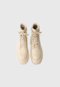 Bottes chelsea en cuir beige avec une finition lisse, bout rond, poignet rembourré et accents en métal doré. Équipées de lacets et d'une fermeture éclair sur le côté.