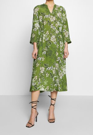 Robe midi verte à imprimé floral blanc, col en V, manches trois-quarts, portée avec des sandales noires à talons hauts à fines lanières.
