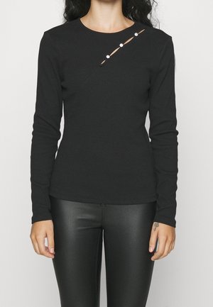 Kobieta ubrana w czarną bluzkę z długim rękawem, z ukośnym wycięciem i trzema ozdobnymi guzikami przy ramieniu, w połączeniu z czarnymi skórzanymi spodniami.